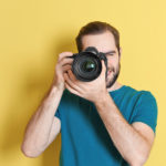 Photographer for Ocean County web deisgn firm with camera up to eye, wearing blue shirt, standing in front of yellow wall